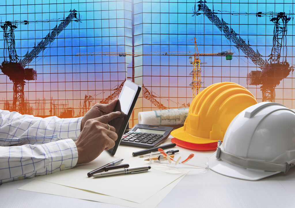 Image of hardhats, documents, and a man holding a tablet at a desk